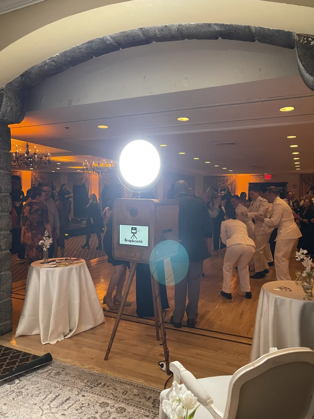 Photo of a group of people dancing at a party or wedding reception, with a photo booth in the foreground and tables with white tablecloths, flowers, and a bright white light in the background.