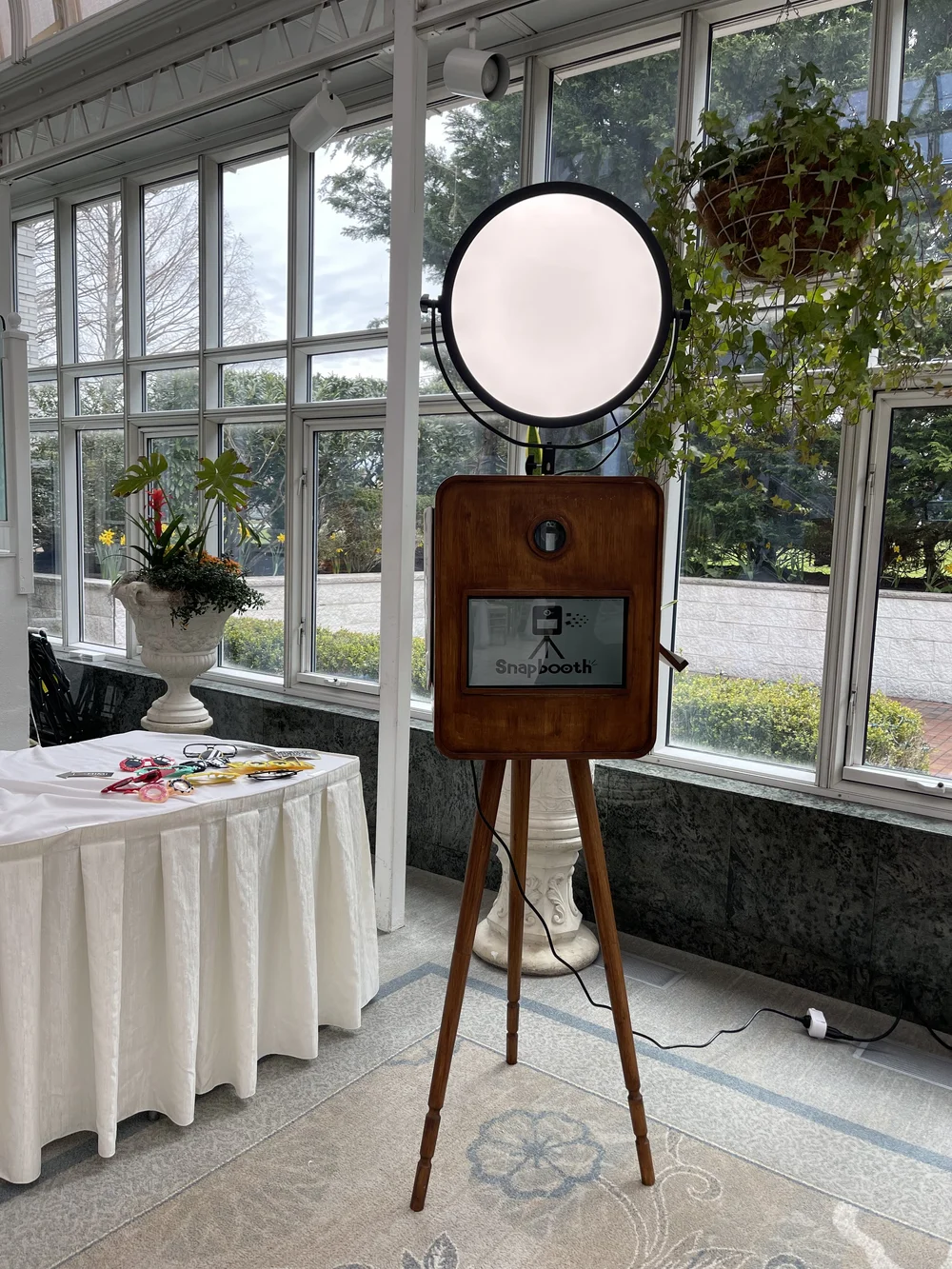 A photo booth setup with a large round light, a wooden-framed screen on a tripod, inside a bright room with large windows and plants outside.