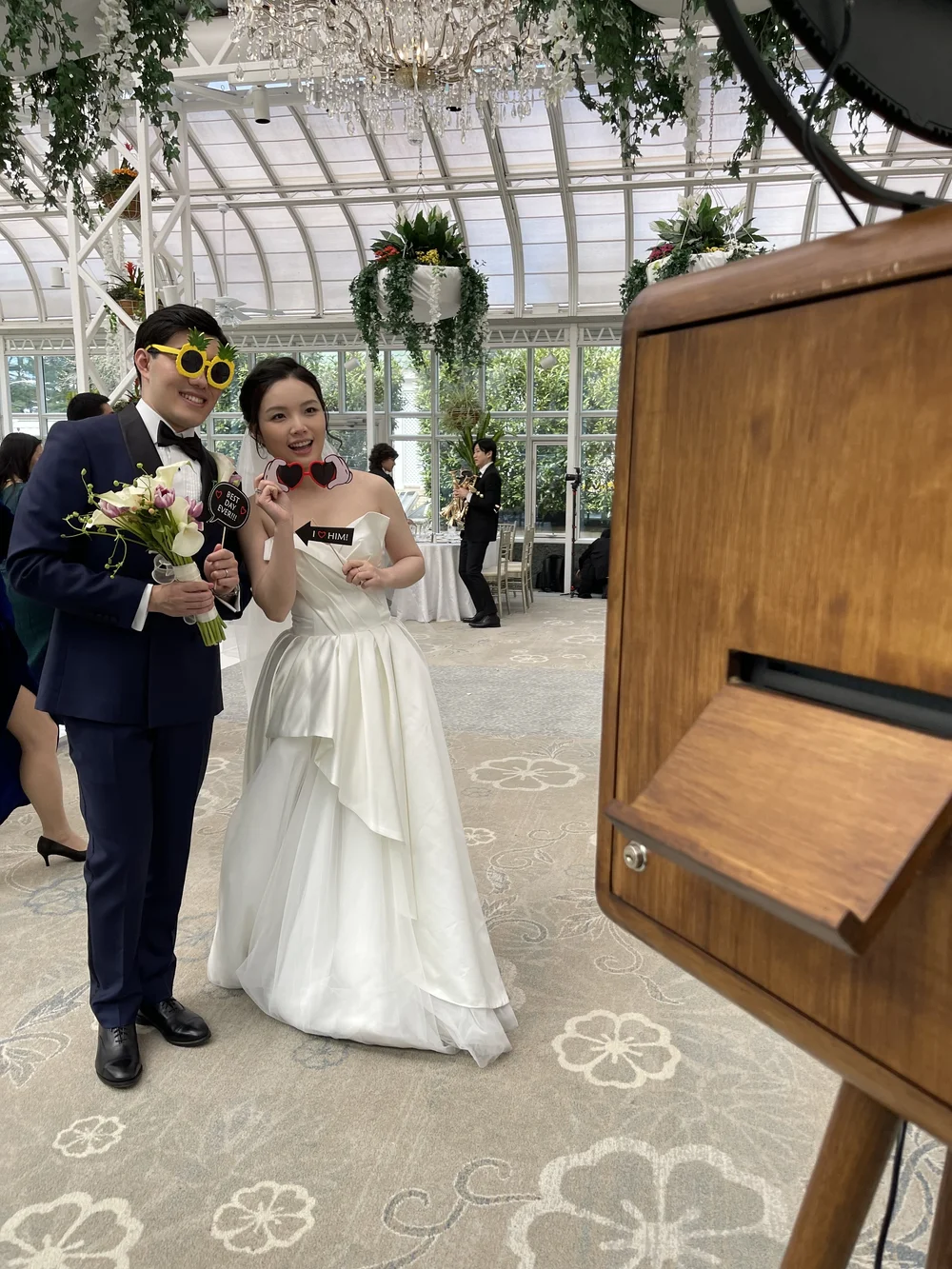 A bride and groom pose for a photo at a wedding reception in a glass conservatory. They wear playful photo props; the groom has pineapple sunglasses, and the bride has red heart-shaped glasses. The bride is in a white strapless wedding gown, and the 