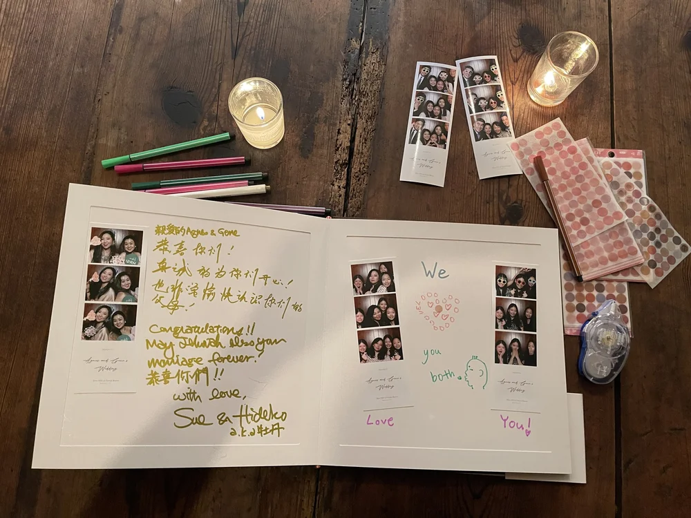 A wedding guestbook with photos, handwritten notes, and decorative drawings, surrounded by photo booth strips, markers, candles, and adhesive dots on a wooden table.