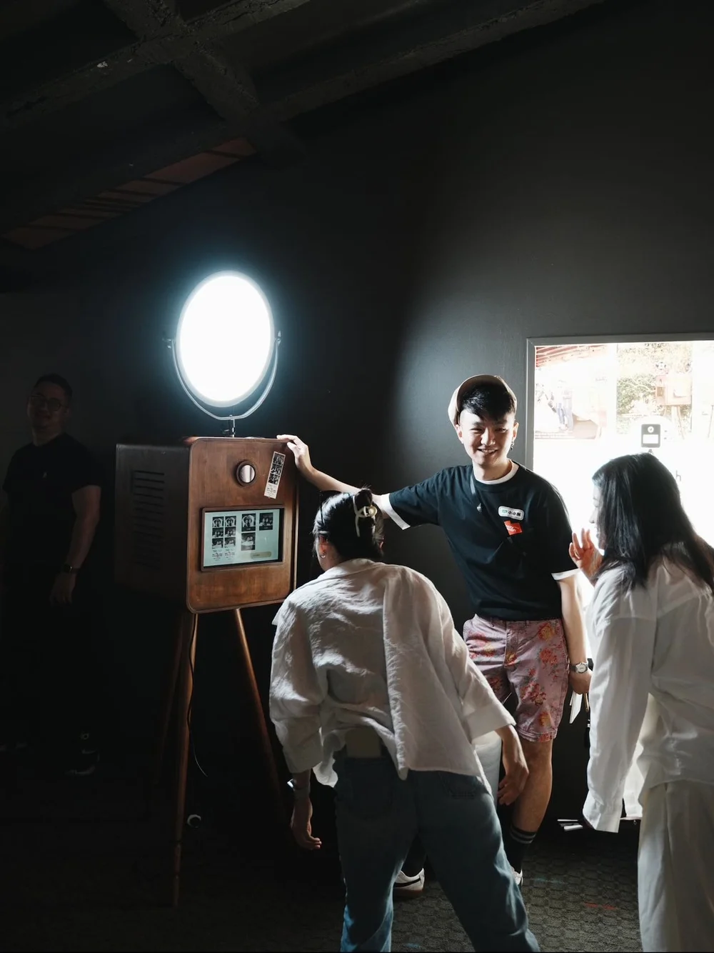 People gathered around a photo booth with an illuminated backdrop, smiling and engaging with each other, in a dimly lit room.