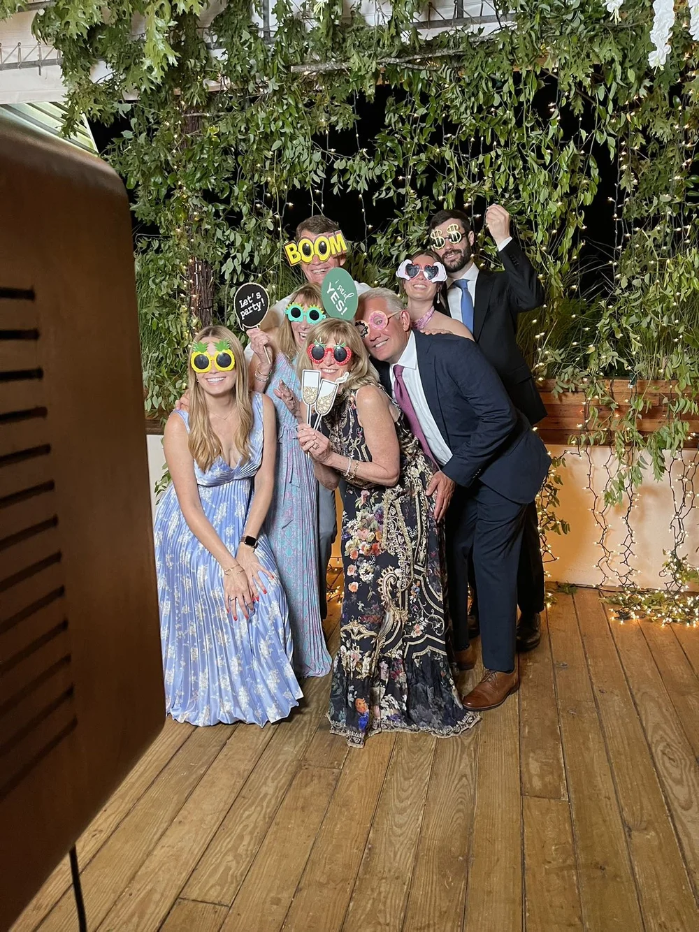 Group of people at a celebration wearing colorful props and holding signs, posing for a photo in front of a leafy green backdrop with fairy lights.