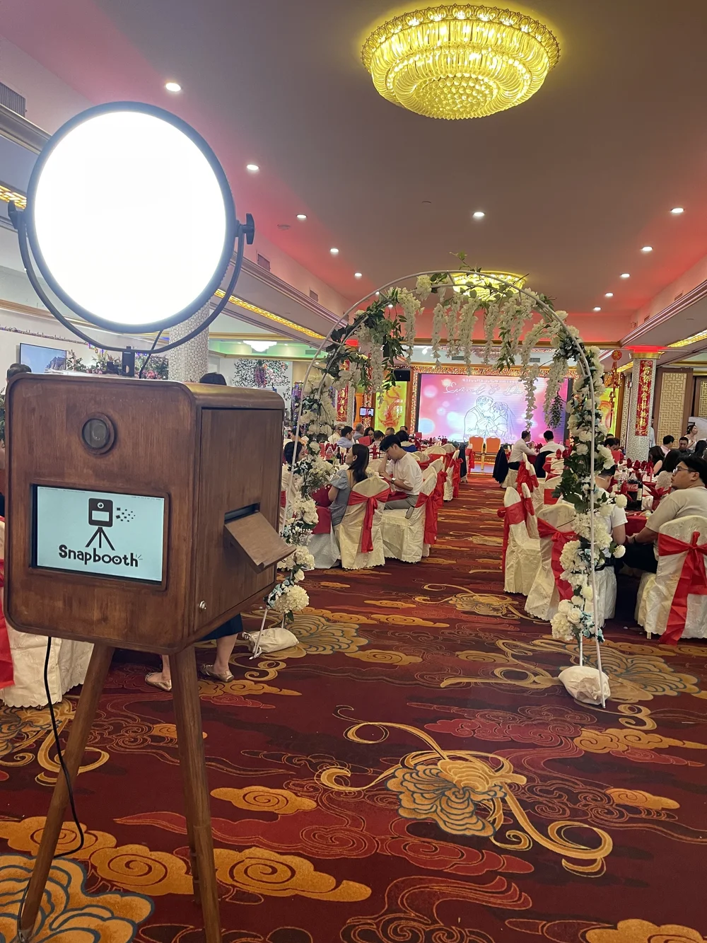 A wedding reception hall decorated with white and red chairs, floral arches, and a stage with a large screen. Guest tables are arranged with chairs draped in white covers and red ribbons.