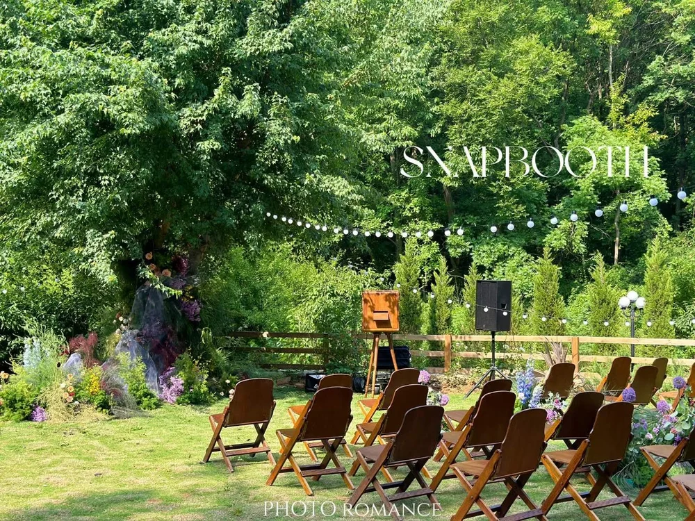 Outdoor wedding ceremony setup with empty wooden chairs, a floral arch, and string lights on a sunny day in a lush green garden.