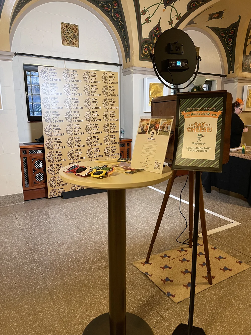Photo booth area at the New York City Center with a sign that says, "Say Cheese!" and a complimentary photo booth. There are colorful sunglasses on a table, a backdrop with the New York City Center logo, a stand with a built-in camera, and a person s