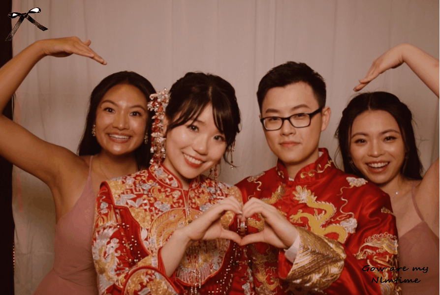 Four people celebrating in traditional Chinese wedding attire, making a heart shape with their hands, smiling, with two women in pink dresses and two men in red embroidered robes, with a white background.